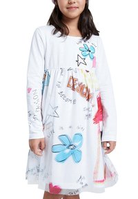Fille portant une robe blanche à manches longues avec des motifs colorés de fleurs et d'étoiles ainsi qu'un texte de style manuscrit, debout devant un fond blanc.