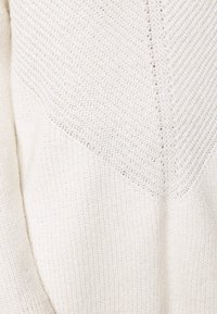 Jasno beżowy sweter dzianinowy z teksturowanym wzorem. Posiada wzór w zygzak na górnej części i gładki splot w dolnej sekcji.