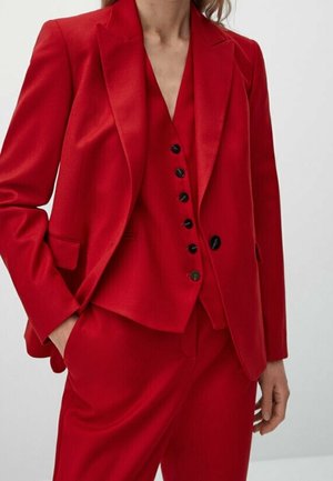 Femme portant un tailleur rouge vif sur mesure avec un blazer, un gilet boutonné et un pantalon assorti, debout avec une main dans la poche.