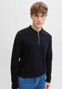 Jauns vīrietis ar gaiši brūniem matiem, tērpies melnā garā piedurkņu adījumā ar priekšējo rāvējslēdzēju un zila džinsa biksēs, uz vienkārša fona.