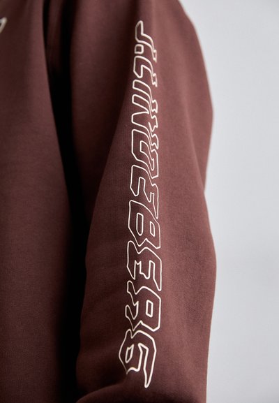 Καφέ φούτερ με λευκό εκτυπωμένο κείμενο κατά μήκος του μανικιού, με απαλή υφή και σχεδιασμό χαλαρής εφαρμογής.