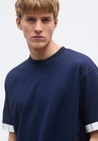 Tmavomodré bavlnené tričko s okrúhlym výstrihom, krátkymi rukávmi a bielymi manžetami s modrými prúžkami. Jednoduchý, pohodlný strih.