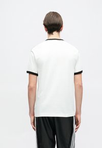 Biela tričko s čiernym rebrovým golierom a manžetami na rukávoch, mäkká textúra, štandardný strih, zobrazené zozadu, v kombinácii s čiernymi teplákmi.