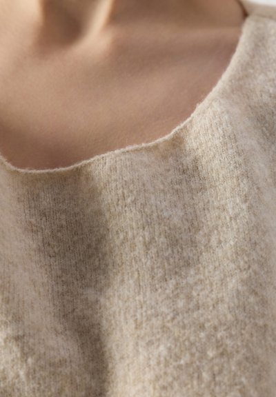Gros plan sur le cou et la clavicule d'une personne portant un pull en tricot beige et doux avec un large col arrondi.