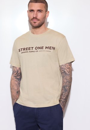 Mand med kort hår og tatoveringer på begge arme iført en beige T-shirt med teksten "STREET ONE MEN" og marineblå bukser står op ad en hvid væg.