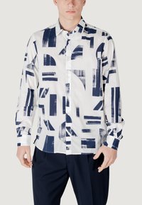 Langarmshirt van witte stof met blauwe abstracte geometrische patronen. Voorzien van een knoopsluiting aan de voorzijde en een standaard kraag.