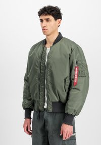 Zeleni bomber jakna napravljena od sjajnog najlona s rebrastim manžetama i rubom, zatvaranjem na zip, te bočnom džepu s detaljem crvene oznake.