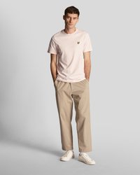 Ljusrosa bomullst-shirt med en liten gul logotyp, i kombination med beige avslappnade byxor och vita sneakers. Enkel, avslappnad design.