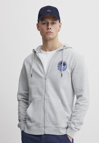 Un sweat à capuche gris zippé avec une poche frontale, arborant un logo bleu rond. Associé à une casquette marine, porté avec un T-shirt blanc en dessous.