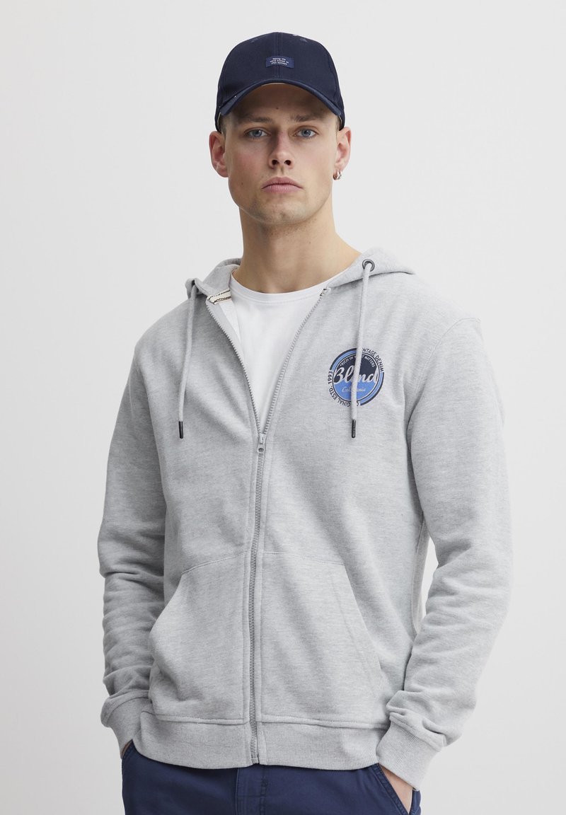 Un sweat à capuche gris zippé avec une poche frontale, arborant un logo bleu rond. Associé à une casquette marine, porté avec un T-shirt blanc en dessous.