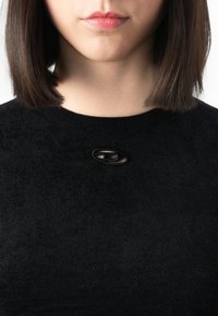 Donna con capelli lisci e scuri che indossa un top nero con un piccolo accessorio ovale nero vicino allo scollo, su uno sfondo bianco.