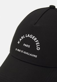 Melnā cepure ar izliekta vizieri, kurā ir balts teksts: "KARL LAGERFELD PARIS 21 RUE ST-GUILAUME." Teksts ir centrāli novietots priekšpusē.