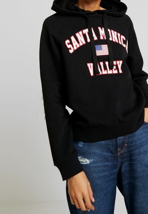 Osoba ubrana w czarną bluzę z kapturem z napisem „Santa Monica Valley” i amerykańską flagą, w parze z niebieskimi podartymi dżinsami, na jednolitym tle.
