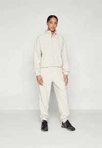 Completo tuta in fleece beige chiaro con pullover a zip quarti e pantaloni con vita elastica, abbinato a scarpe sportive nere. Texture liscia.