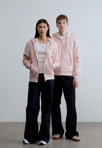 Δύο μοντέλα στέκονται δίπλα-δίπλα φορώντας ταιριαστά ανοιχτό ροζ φούτερ με φερμουάρ και κουκούλα. Τα φούτερ έχουν λογότυπο στο στήθος. Και οι δύο φορούν σκούρα τζιν και casual παπούτσια.