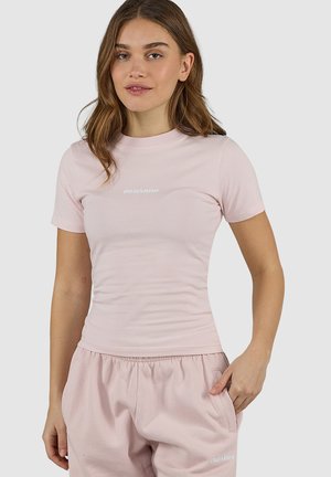 Młoda kobieta ubrana w dopasowany, jasnoróżowy T-shirt i pasujące, luźne szorty, stojąca z jedną ręką w kieszeni na jednolitym tle.