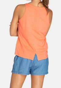 Top senza maniche arancione con design a schiena aperta, abbinato a pantaloni corti in denim blu. Materiale in cotone misto, texture leggera.