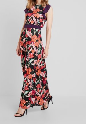 Vrouw die een zwarte maxi-jurk draagt met oranje en rode bloemenprint, paarse accenten en zwarte sandalen met hoge hakken, staand op een grijze achtergrond.
