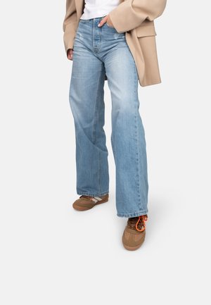 Jean large bleu clair avec une finition délavée, associé à des chaussures marron. La tenue est complétée par un blazer beige.
