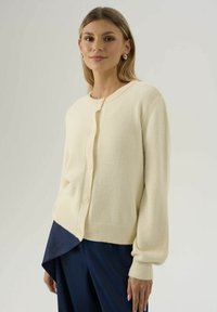 Donna che indossa un cardigan cream con bottoni e maniche lunghe, abbinato a pantaloni asimmetrici blu navy, mentre sta in piedi davanti a uno sfondo semplice.