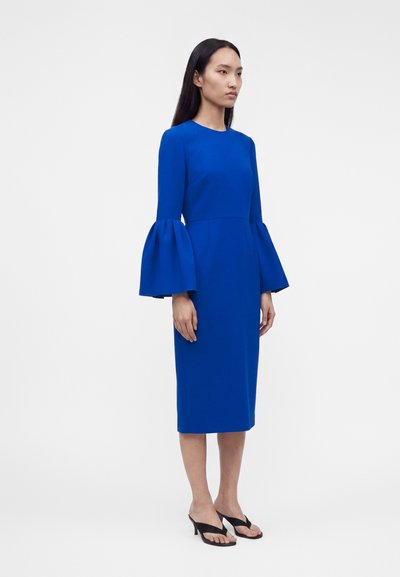 Γυναίκα στέκεται, φοράει ένα γονατο-length φόρεμα σε βασιλικό μπλε με μακριά, μαζεμένα μανίκια καμπάνα και μαύρες ανοιχτές, ψηλοτάκουνες σανδάλες.