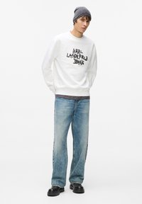 Bijela majica dugih rukava s natpisom "KARL LAGERFELD JEANS" ispisanim crnim slovima, uparena s ispucalim plavim trapericama i crnim debelim cipelama. Na glavi nosi sivi vuneni šešir.