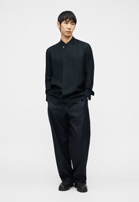 Uomo con capelli neri che indossa una camicia nera con collo alto e pantaloni larghi a righe sottili scuri, in piedi con le mani in tasca.