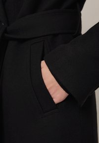 Czarny płaszcz o gładkiej fakturze, z boczną kieszenią i detalem paska. Materiał wygląda na miękki i lekko teksturowany.