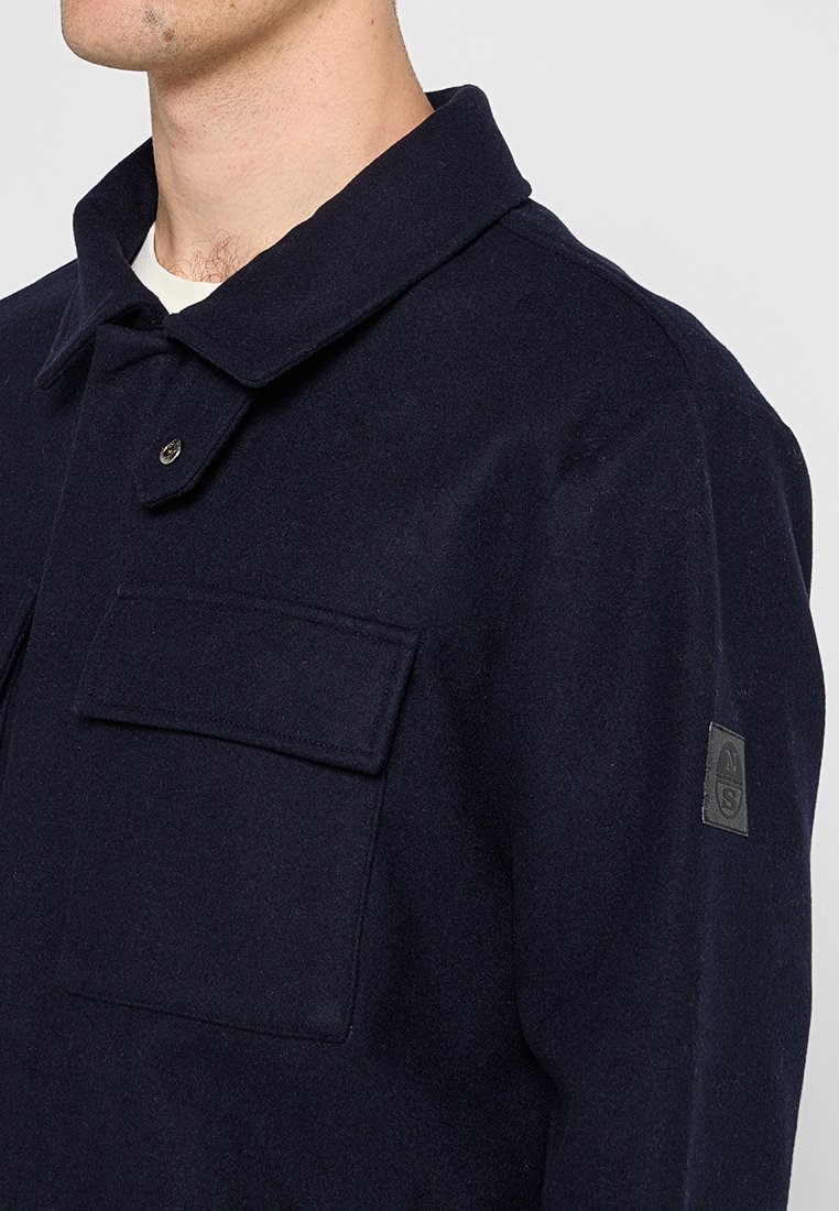 Dunkelblaues Wolljackett mit Kragen, einer einzelnen Brusttasche und Seitennähten. Es zeichnet sich durch einen strukturierten Stoff und ein Logopatch am Ärmels aus.