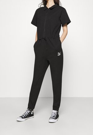 Sort kortærmet jumpsuit med lynlåsfront, snøre talje og logo detaljer. Blødt stof med afslappet pasform, parret med sorte sneakers.