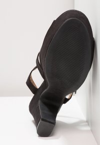 Chaussure à talon haut en suède noir avec un talon courbé, un design ouvert et une semelle en caoutchouc texturé comportant des rainures horizontales.