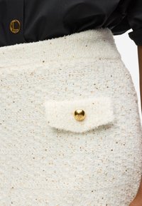 Fehér textúrájú szoknya arany flitterekkel, kiemelkedő aranygombbal a hajtókás zsebnél. Testre szabott fazon magas derékkal.