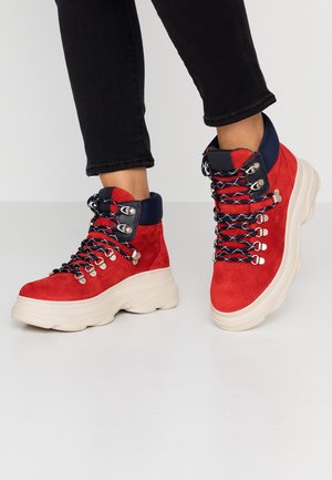 Rode suède hoge sneakers met marineblauwe gewatteerde kragen, zwart-witte veters en dikke witte rubberen zolen, gedragen met zwarte broek.