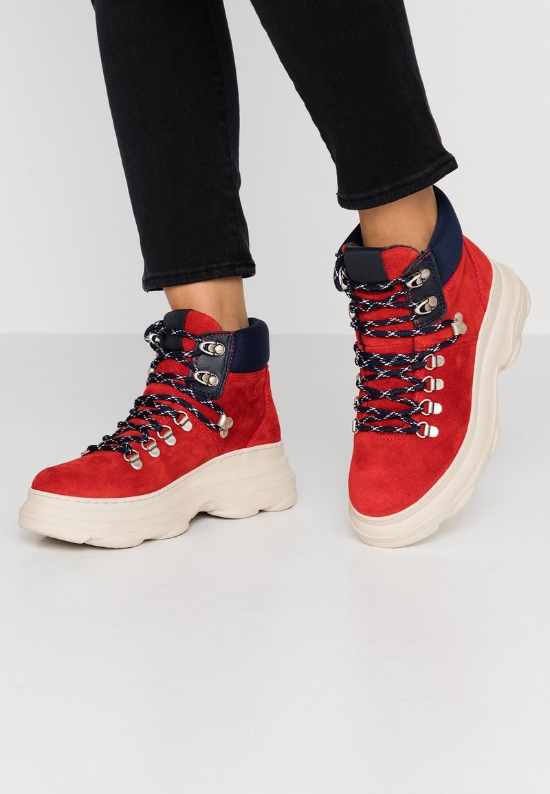 Baskets montantes en daim rouge avec col en nylon rembourré bleu marine, lacets noirs et blancs, et semelles épaisses en caoutchouc blanc, portées avec un pantalon noir.