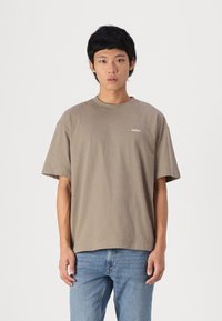 Bruine katoenen T-shirt met een ronde hals, korte mouwen en een klein wit logo op de linkerkant van de borst. Gepaard met lichtblauwe jeans.