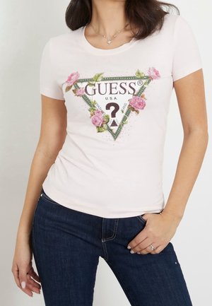 Kvinde iført en lyserød Guess t-shirt med en blomsterprint trekantet logo, kombineret med mørkeblå jeans, stående med den ene hånd i lommen.