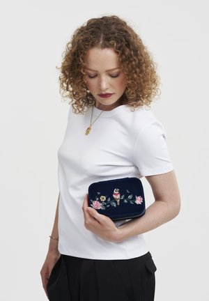 Mornarska žametna torbica z vezenjem cvetja in ptic, pravokotne oblike, gladke teksture in zadrgo. Držana ob beli srajci.