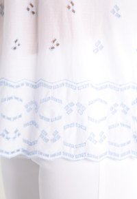Hvid stofoberk med lyseblå broderede diamanter og geometriske mønstre, med en dekorativ blonde-lignende kant. Glat tekstur.