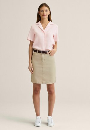 Kvinde stående iført en lyserød kortærmet skjorte med knapper, beige knælængde nederdel med bælte og hvide sneakers på en ensfarvet baggrund.