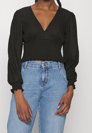 Žena nosící černý texturovaný top s dlouhým rukávem a výstřihem do V, s řaseným pasem a světlými modrými džíny s vysokým pasem, stojící před jednobarevným pozadím.