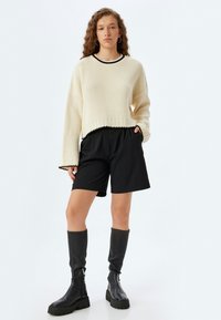 Maglione a maglia con texture cremosa e finiture nere, abbinato a pantaloni corti neri su misura. Il modello indossa stivali neri fino al ginocchio con suole spesse.