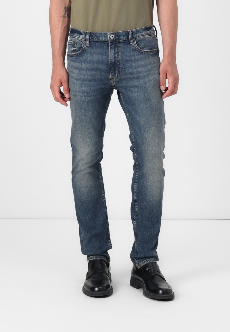 Vaqueros de denim azul con corte recto, que presentan áreas desvaídas, diseño de cinco bolsillos y cierre de botón de metal. Combinados con zapatos negros.