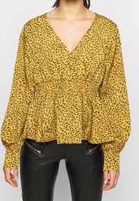 Gul blus med leopardmönster, djup V-ringning och puffärmar. Har elastisk midja och flytande peplumslits. Kombinerad med svarta läderbyxor.