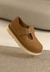 Bruine suède schoen met een ronde neus, uitsnijding aan de voorkant en een klittenbandsluiting. De zool is lichtgeel en gestructureerd voor grip.