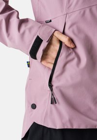 Lila jacka med en figursydd design, som har en dragkedjeficka, justering vid ärmslut med kardborre och en svart knappdetalj längst ner.