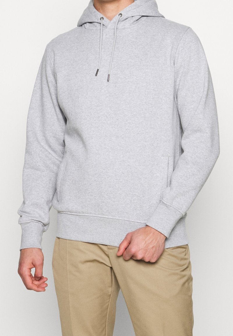 Sudadera de algodón gris con capucha ajustable, bolsillo canguro en la parte delantera, puños acanalados y dobladillo. Lleva puesta sobre pantalones beige.