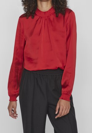 Femme portant un chemisier rouge à manches longues avec poignets et encolure froncés, rentrée dans un pantalon noir taille haute à taille élastique.