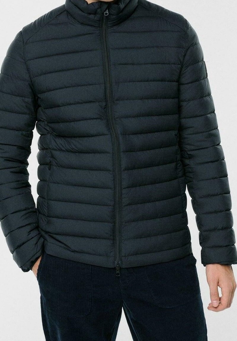 Černá vycpaná bunda s vysokým límcem, vodorovným prošíváním a předním zipem. Nošena s tmavými manšestrovými kalhotami.