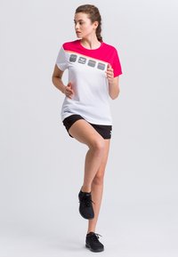 T-shirt sportivo bianco e rosa con maniche corte, caratterizzato da un logo e quattro pannelli decorativi sul petto; abbinato a pantaloni corti neri e sneakers.