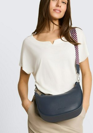 Donna con top bianco e pantaloni beige che porta una borsa a tracolla blu navy con una tracolla decorata sulla spalla.